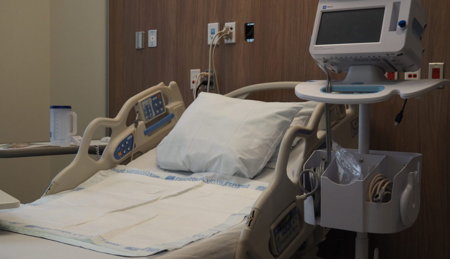 A view of a vacant hospital bed with monitoring equipment on a stand next to the bed.