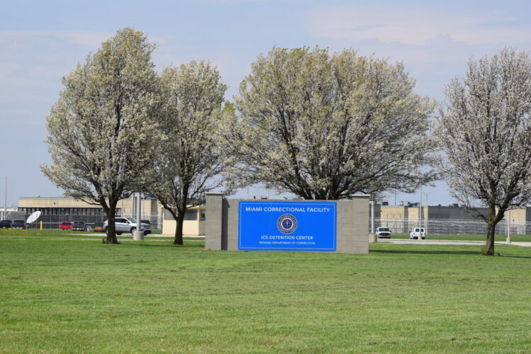 A blue sign reads Miami Correctional Facility. Behind it, through a screen of trees, the prison building can be seen.