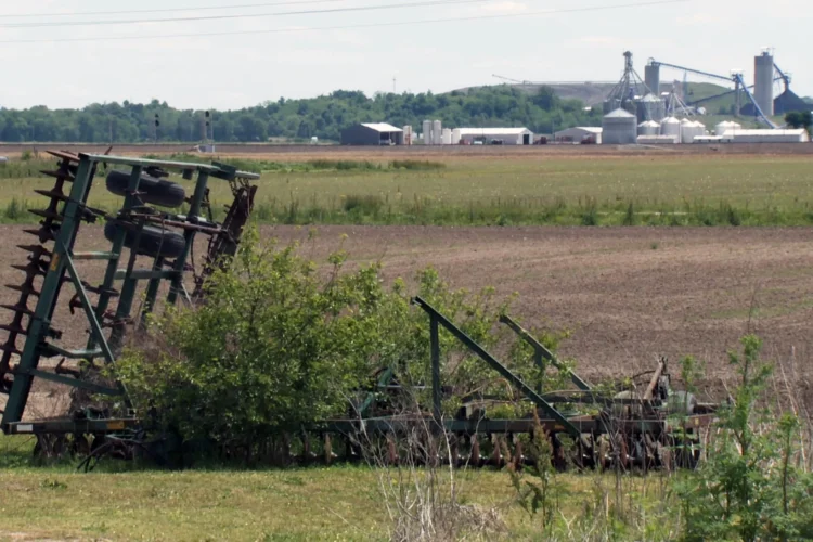 A large farm field is pictured with a huge complex of marns and grain bins in the far background. In the foreground is an abandoned piece of farm machinery with weeds and small trees growing through it.