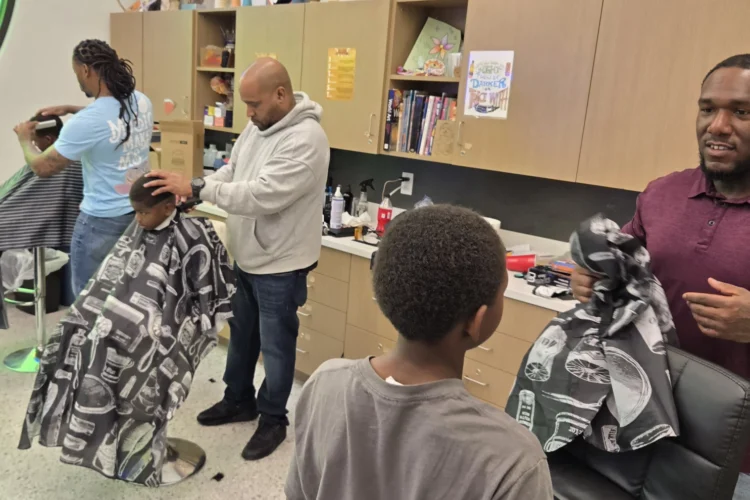 A photograph in a barbershop with three adult barbers giving three boys haircuts.