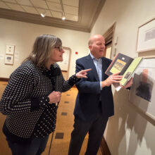 Michelle Kinsey and Robert G. LaFrance look at framed artwork on a wall. Robert is holding up a book to one of the framed pieces.