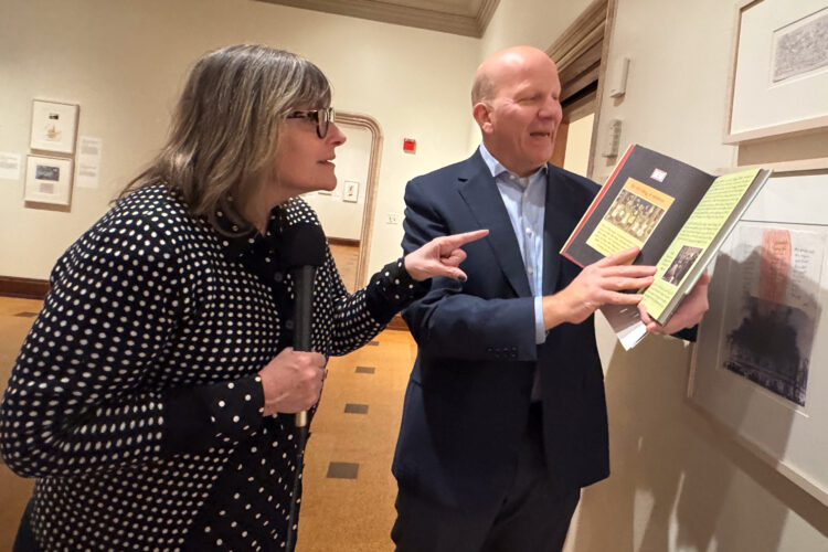 Michelle Kinsey and Robert G. LaFrance look at framed artwork on a wall. Robert is holding up a book to one of the framed pieces.