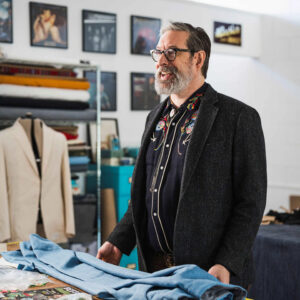 Jerry Lee Atwood smiles in a workshop filled with cloth and thread. He is wearing his brand Union Western, with an embroidered shirt.