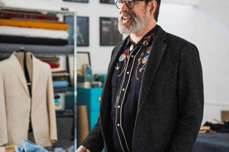 Jerry Lee Atwood smiles in a workshop filled with cloth and thread. He is wearing his brand Union Western, with an embroidered shirt.