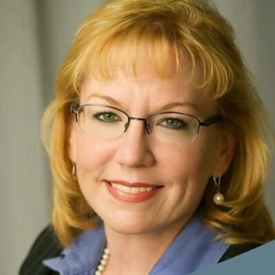 Joann McKinney poses for a headshot. She has blond hair and wears glasses with a blue collared shirt and black jacket.