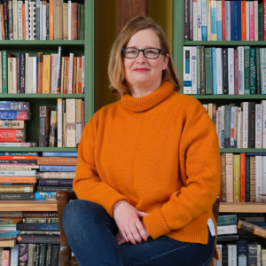 Lucy Mangan sits in front of bookshelves. She is a white woman with shoulder-length strawberry blonde hair. She is wearing an orange turtle-neck sweater, blue jeans, red lipstick, and black glasses.