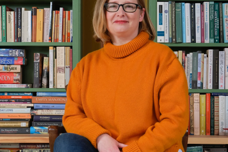 Lucy Mangan sits in front of bookshelves. She is a white woman with shoulder-length strawberry blonde hair. She is wearing an orange turtle-neck sweater, blue jeans, red lipstick, and black glasses.