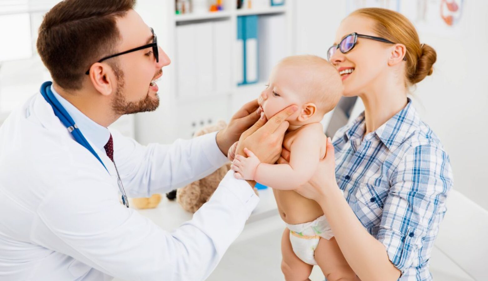 An infant baby is held up by his mother as a doctor in a white coat and glasses cups its chin, looking into its eyes.
