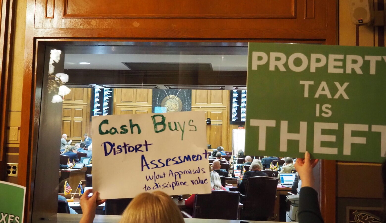 Protesters hold signs outside the Indiana Statehouse chambers. One reads Property Tax is Theft. Another reads Cash Buys Distort Assessments.