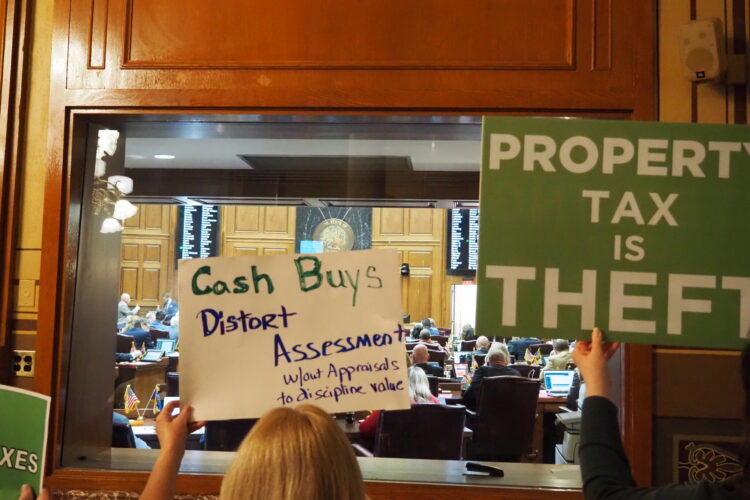 Protesters hold signs outside the Indiana Statehouse chambers. One reads Property Tax is Theft. Another reads Cash Buys Distort Assessments.