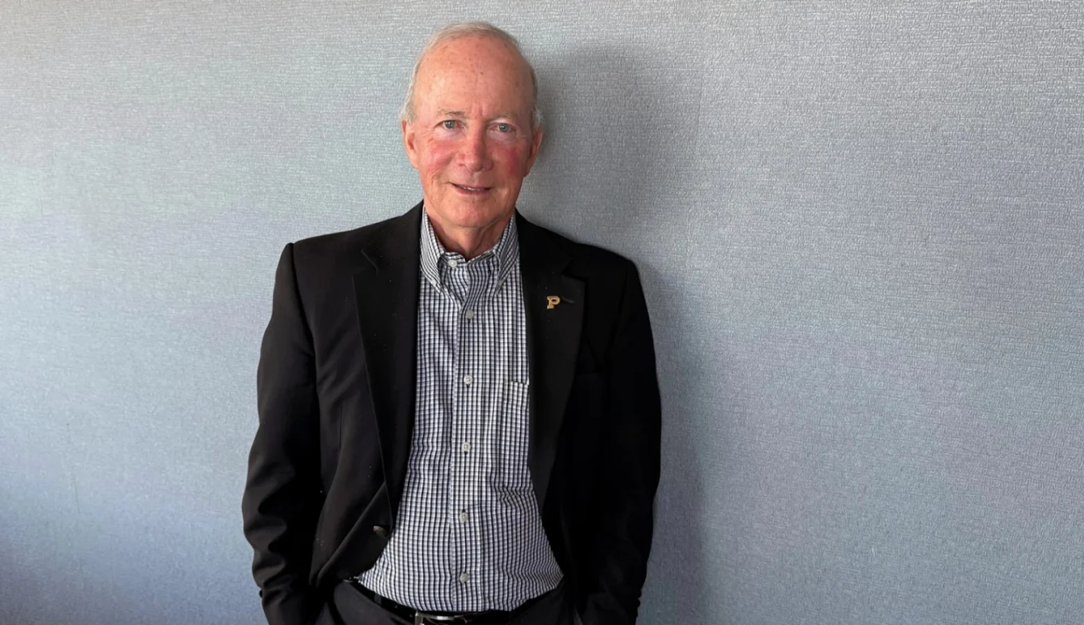 An interior photo of Mitch Daniels wearing a sport coat and checkered shirt without a tie standing against a gray wall.