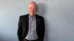 An interior photo of Mitch Daniels wearing a sport coat and checkered shirt without a tie standing against a gray wall.