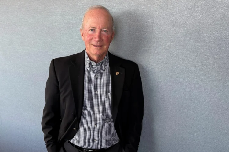 An interior photo of Mitch Daniels wearing a sport coat and checkered shirt without a tie standing against a gray wall.