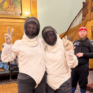 Jen Blackmer and Michelle Kinsey stand in thick white coats with mesh face masks. In the background, Muncie Fencing Club instructor Christian Achgill wears all black and looks on.