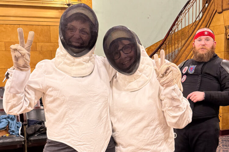 Jen Blackmer and Michelle Kinsey stand in thick white coats with mesh face masks. In the background, Muncie Fencing Club instructor Christian Achgill wears all black and looks on.