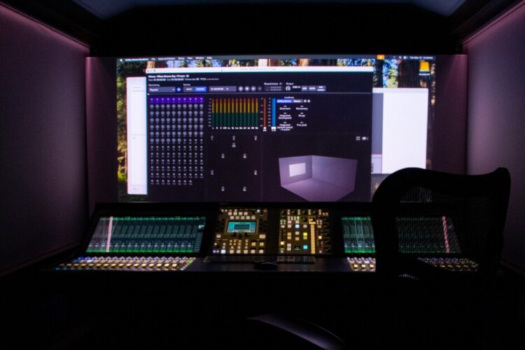 A view inside of the mobile Atmos unit shows a darkened control room with a large video screen in front of a mixing console and a chair.