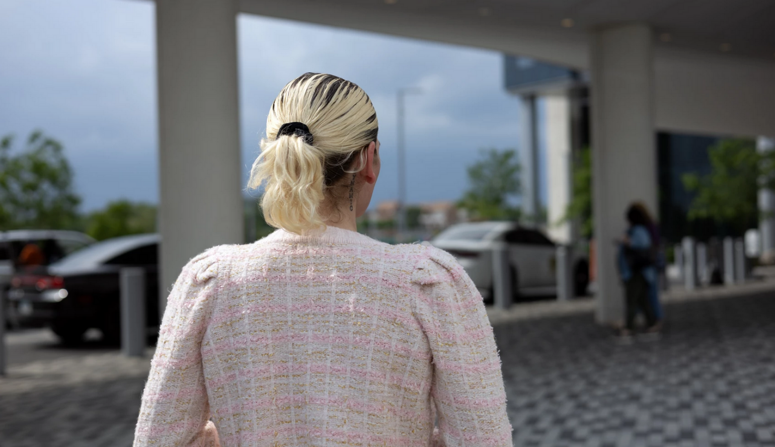 A person, seen from behind, has blond hair and wears a light pink suit jacket.