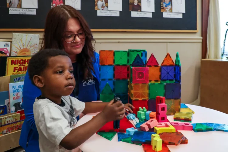 A child and staff member at St. Mary’s Early Childhood Center in Indianapolis. They are seated at a child-height play table with large, colorful, puzzle items with a rack of paperback books behind them.