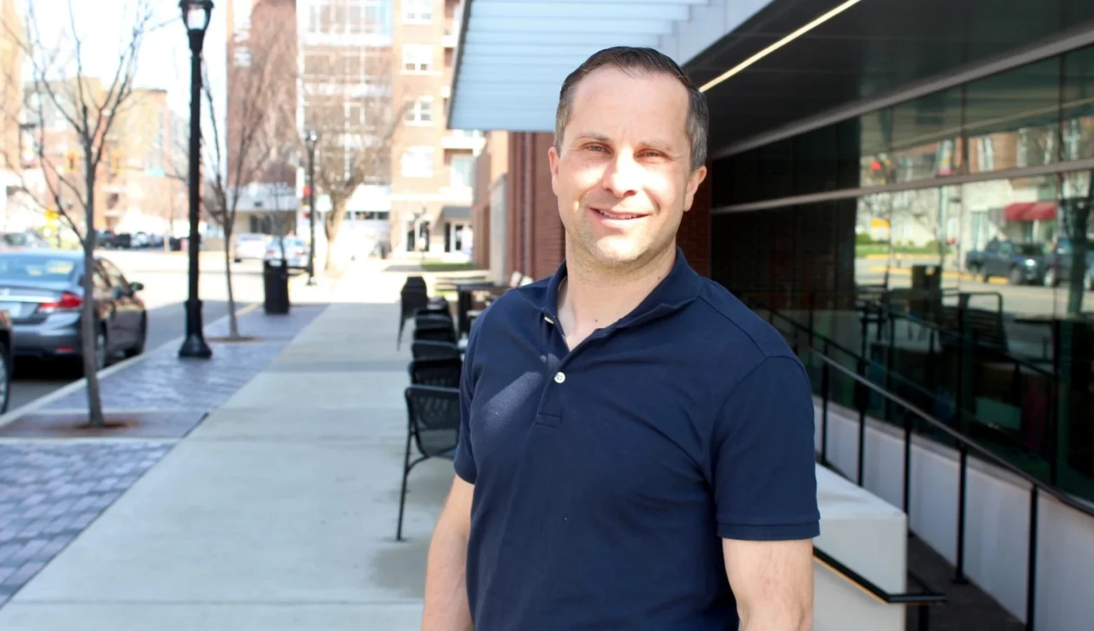 Senator Spencer Deery stands on the sidewalk outside of a building. He faces forward and smiles.