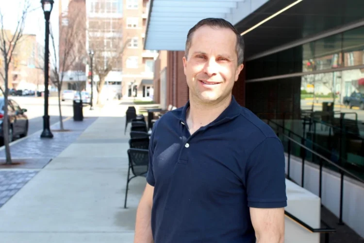 Senator Spencer Deery stands on the sidewalk outside of a building. He faces forward and smiles.