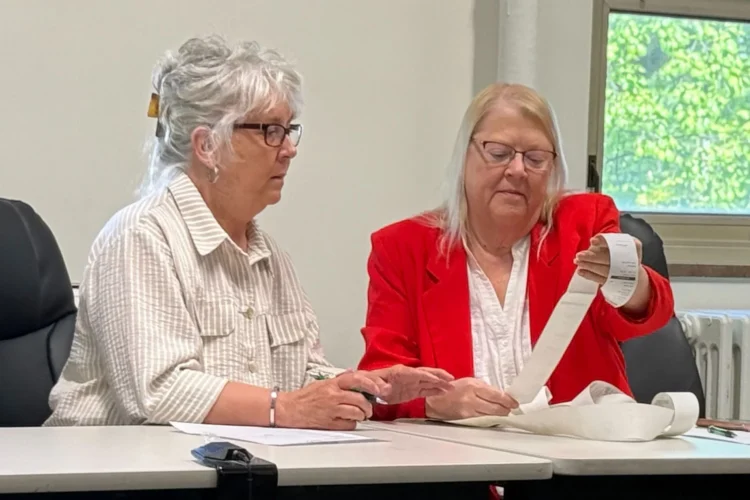 Two women sit at a table and read a long piece of receipt paper from an electronic polling machine.