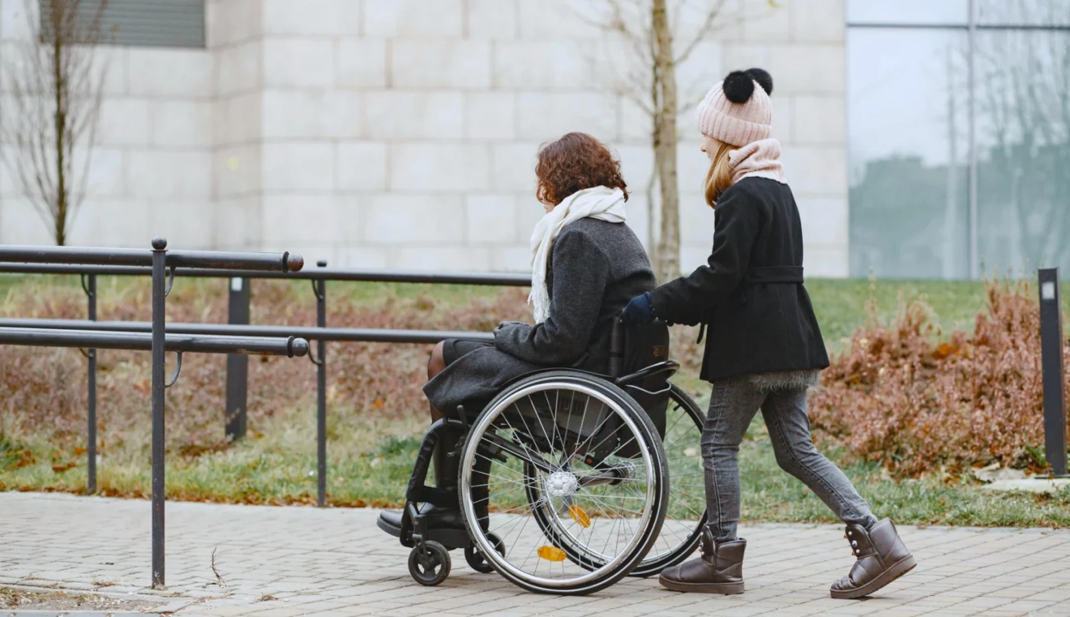 A woman pushes a woman sitting in a wheelchair up an accessible ramp.