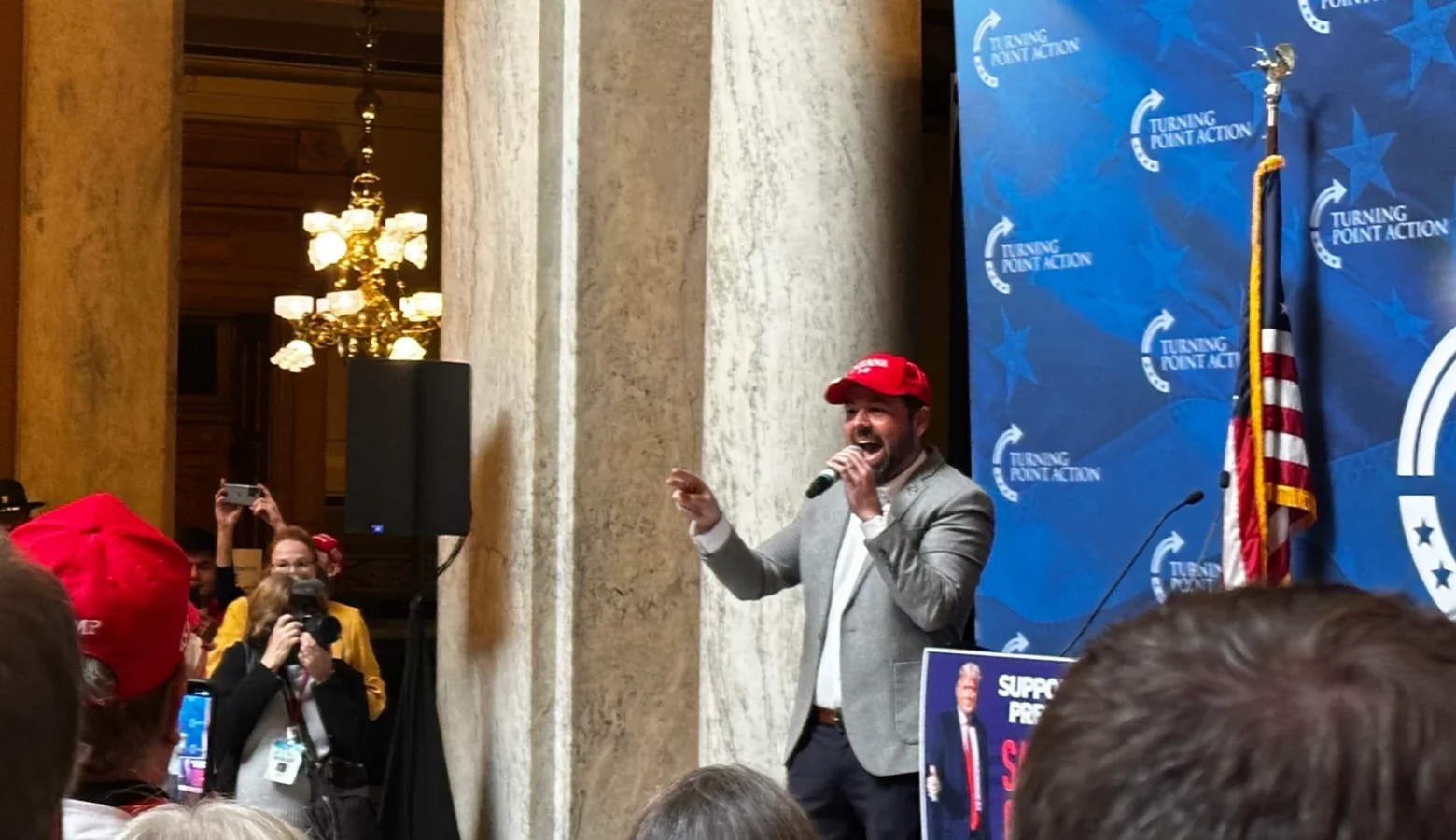 Lieutenant Governor Micah Beckwith stands in the Indiana Statehouse at a Turning Point USA press conference, yelling into a microphone in front of a crowd of people.