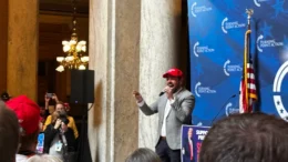 Lieutenant Governor Micah Beckwith stands in the Indiana Statehouse at a Turning Point USA press conference, yelling into a microphone in front of a crowd of people.
