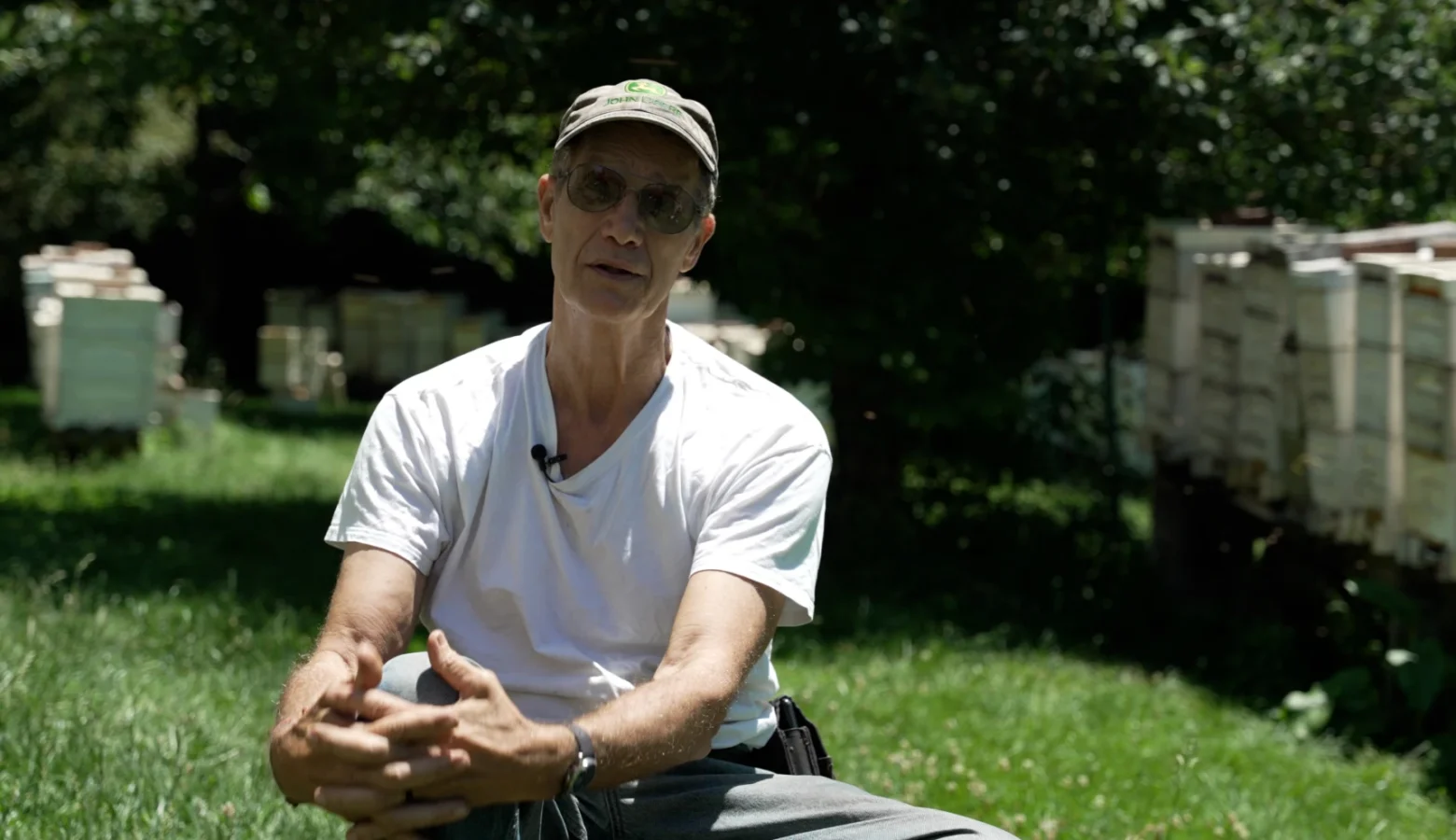 An exterior photo of beekeeper Joe May sitting on a chair in the sunlight in a grassy yard surrounded by trees and bee hives.