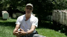 An exterior photo of beekeeper Joe May sitting on a chair in the sunlight in a grassy yard surrounded by trees and bee hives.