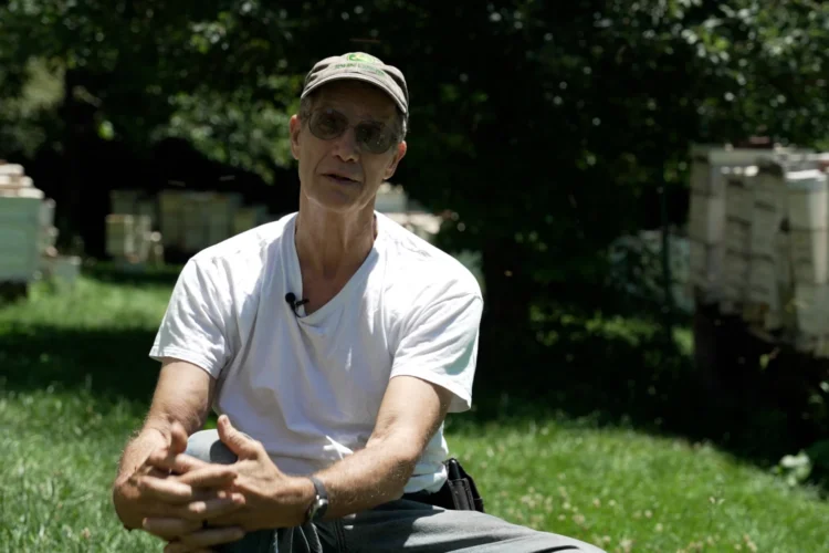 An exterior photo of beekeeper Joe May sitting on a chair in the sunlight in a grassy yard surrounded by trees and bee hives.