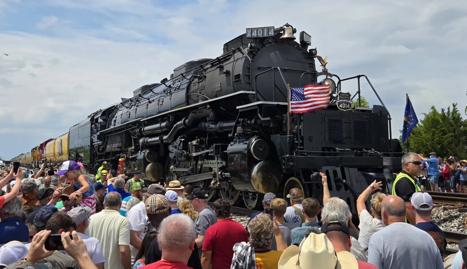 The engine, painted black, is bedecked by an American flag on the left front of the train on a four-foot-tall flagpole; the Indiana state flag is on a similar pole on the right front side of the engine. A large crowd flanks both sides of the train on a sunny day in northern Indiana.