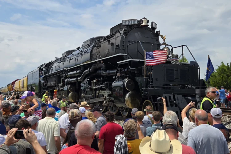 The engine, painted black, is bedecked by an American flag on the left front of the train on a four-foot-tall flagpole; the Indiana state flag is on a similar pole on the right front side of the engine. A large crowd flanks both sides of the train on a sunny day in northern Indiana.