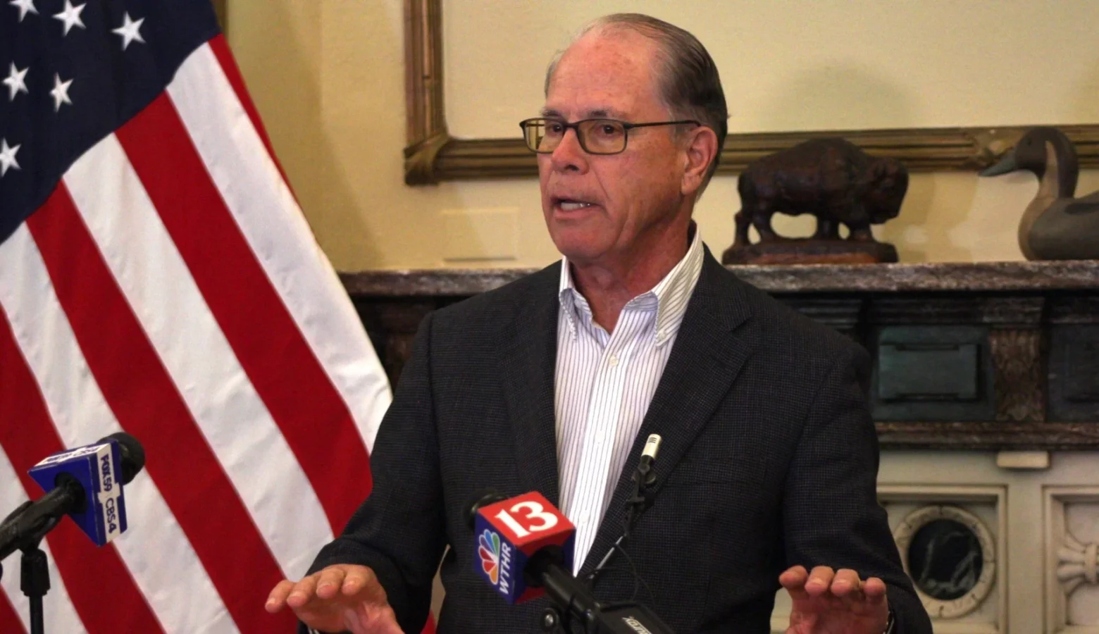 Governor Braun speaks from a podium with TV news microphones visible and an American flag in the left side of the frame.
