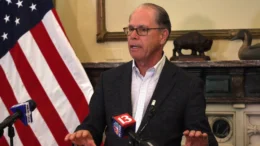 Governor Braun speaks from a podium with TV news microphones visible and an American flag in the left side of the frame.