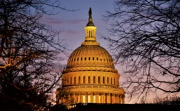 A sunset view of the U.S. Capitol Dome with lighting giving the structure a golden glow. The dome is framed by trees without leaves, presumably in the winter. The sky is clear.