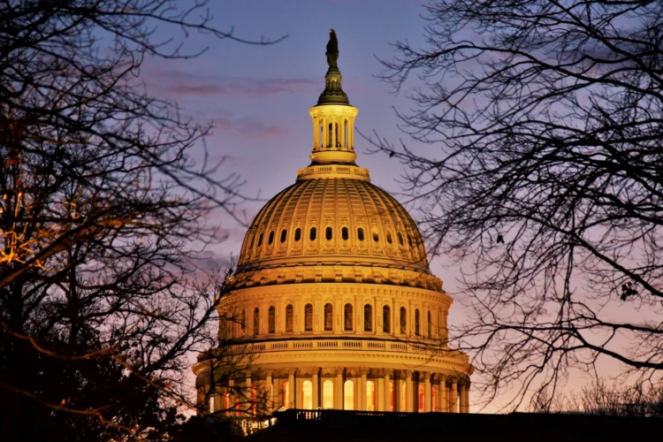 A sunset view of the U.S. Capitol Dome with lighting giving the structure a golden glow. The dome is framed by trees without leaves, presumably in the winter. The sky is clear.