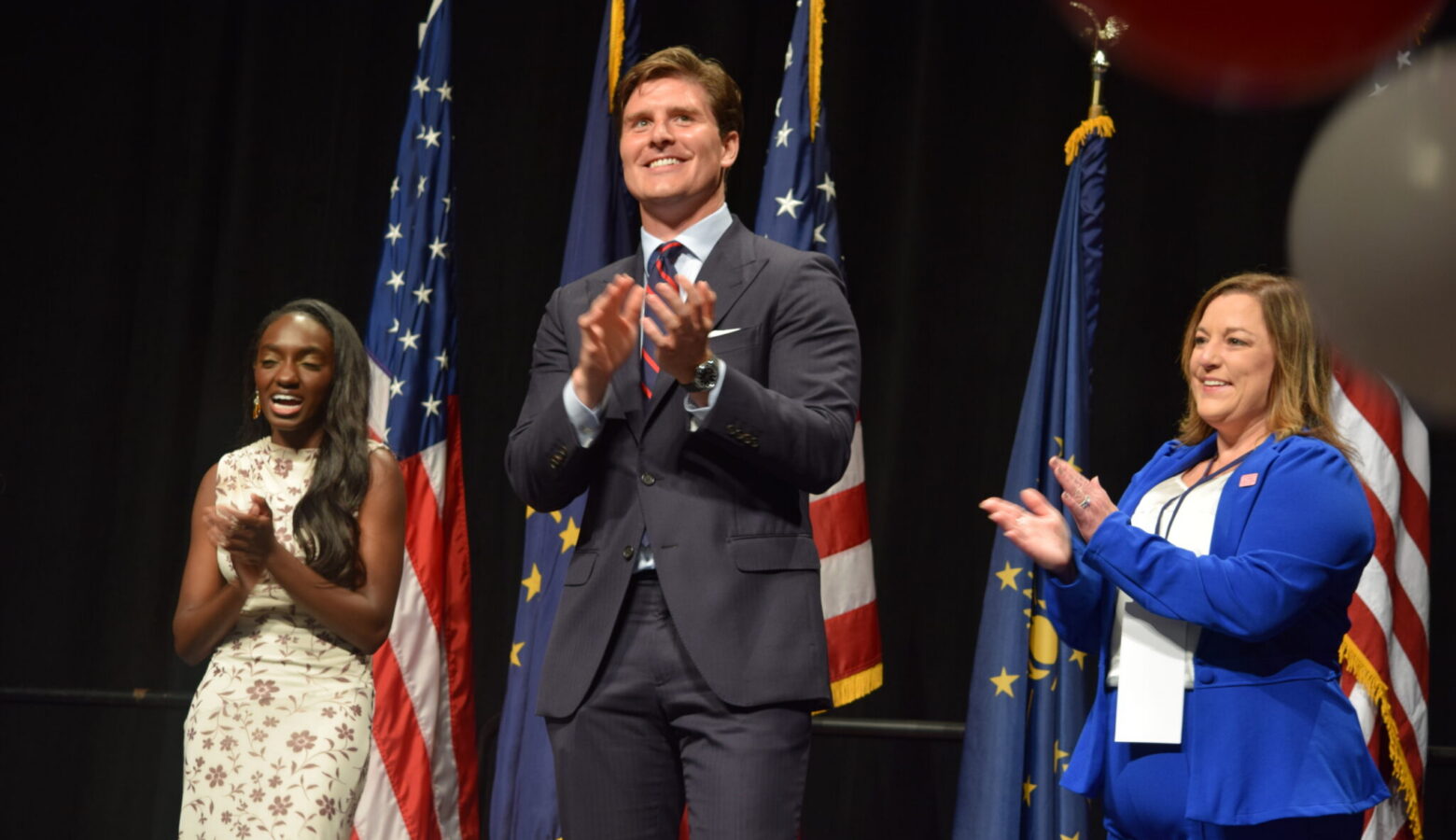 Three Democratic candidates, two women and one man, stand on a stage with several American flags, clapping.