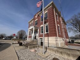 A small brick building with two floors. A flag pole with the american flag stands on the ground just in front of it