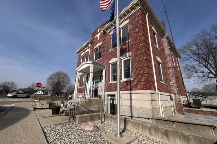 A small brick building with two floors. A flag pole with the american flag stands on the ground just in front of it