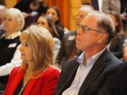Governor Mike Braun and First Lady Maureen Braun sit in a crowd, listening to a speaker at the Indiana Statehouse.