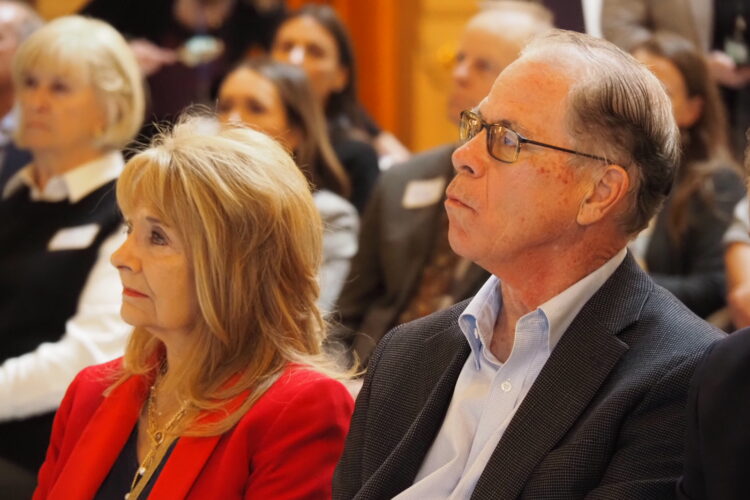 Governor Mike Braun and First Lady Maureen Braun sit in a crowd, listening to a speaker at the Indiana Statehouse.