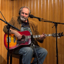 Long Haul Paul sits in front of microphone stands holding a guitar. He is wearing overalls and a brown blazer.