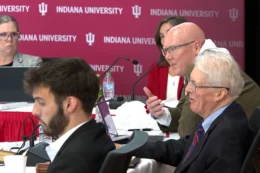 IU trustees are seated at a table conducting business in front of an Indiana University backdrop with the IU logo. ounterclockwise from bottom: Isaac White, James Bopp Jr., J. Timothy Morris, interim secretary Sarah Tosick, and Marilee Springer.