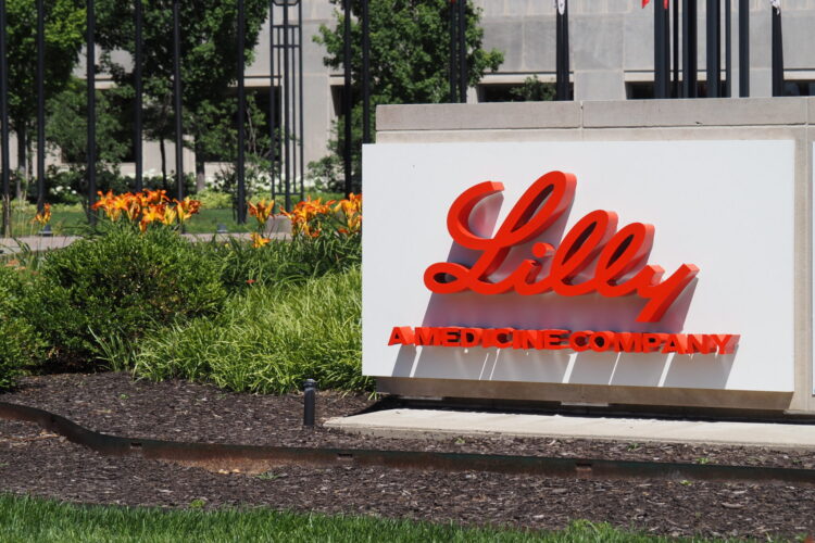 A white sign with red letters displaying the Lilly logo and the phrase "A Medicine Company." The sign is sunlit on a grassy lawn with flowers in the background in front of a large building.