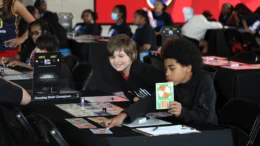 Students Lincoln Schultz and Cameron Coe look through game materials before their first NBA Math Hoops match at Gainbridge Fieldhouse in Indianapolis Tuesday, June, 9, 2026. Other children are seated at nearby tables with computers in front of them along with game cards and sheets with shapes with bright colors.