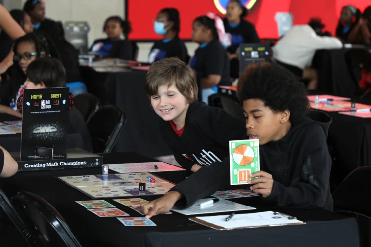 Students Lincoln Schultz and Cameron Coe look through game materials before their first NBA Math Hoops match at Gainbridge Fieldhouse in Indianapolis Tuesday, June, 9, 2026. Other children are seated at nearby tables with computers in front of them along with game cards and sheets with shapes with bright colors.