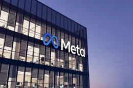 An exterior shot of an office building with the Meta logo featuring a blue loop. The building features mostly windows of glass revealing lit offices within.