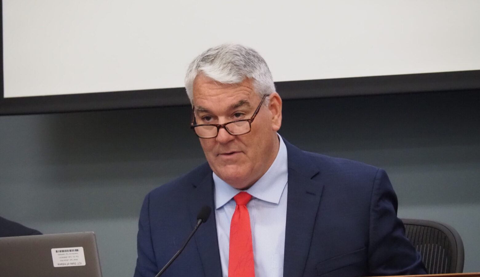 Andy Zay leads a meeting. He is a white man with glasses, white hair, and wearing a blue suit with a red tie.