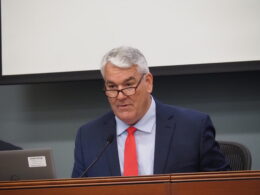 Andy Zay leads a meeting. He is a white man with glasses, white hair, and wearing a blue suit with a red tie.